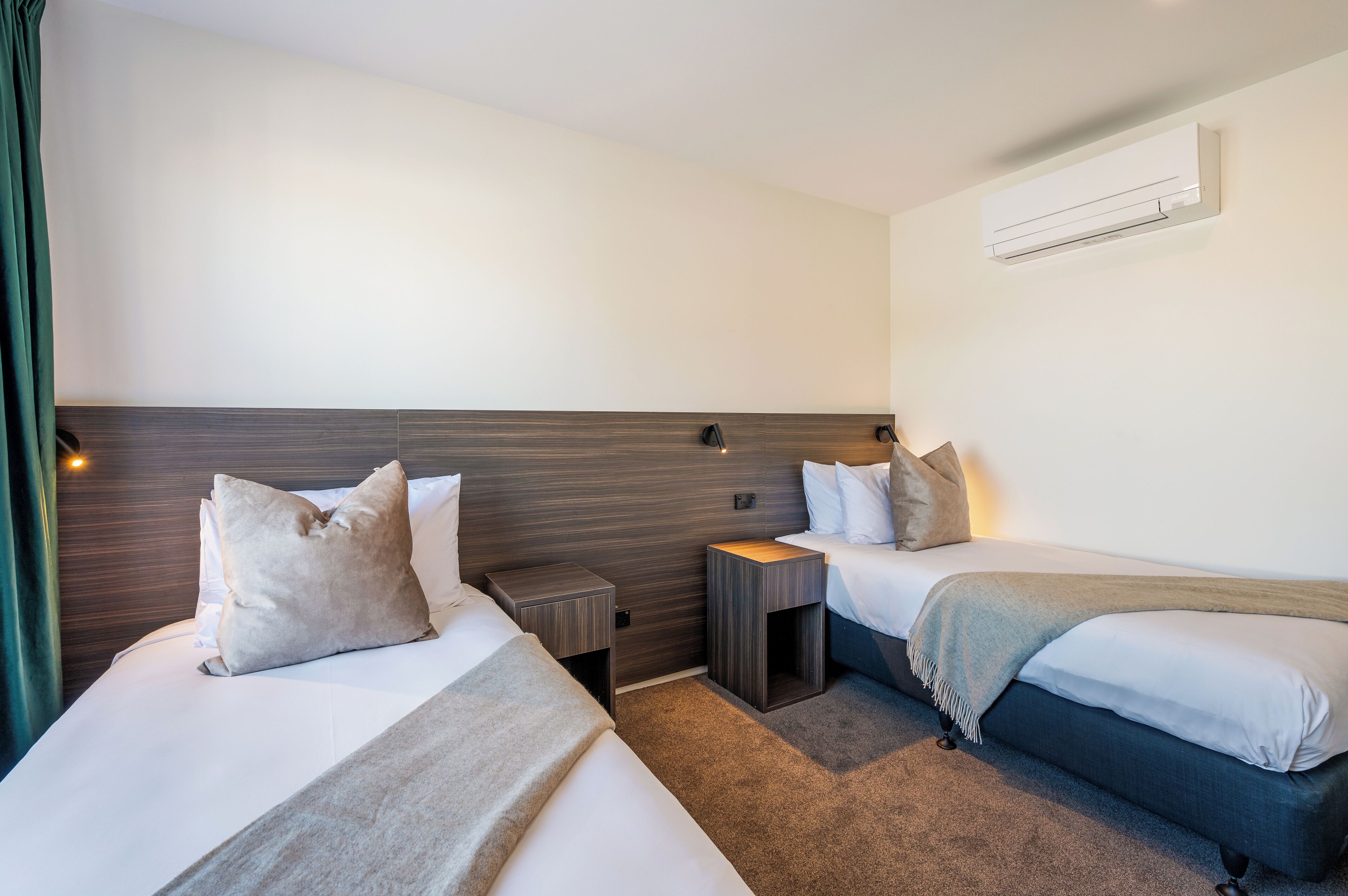A hotel room with two beds, a wooden headboard, a bedside table, a nightstand, and an air conditioning unit.