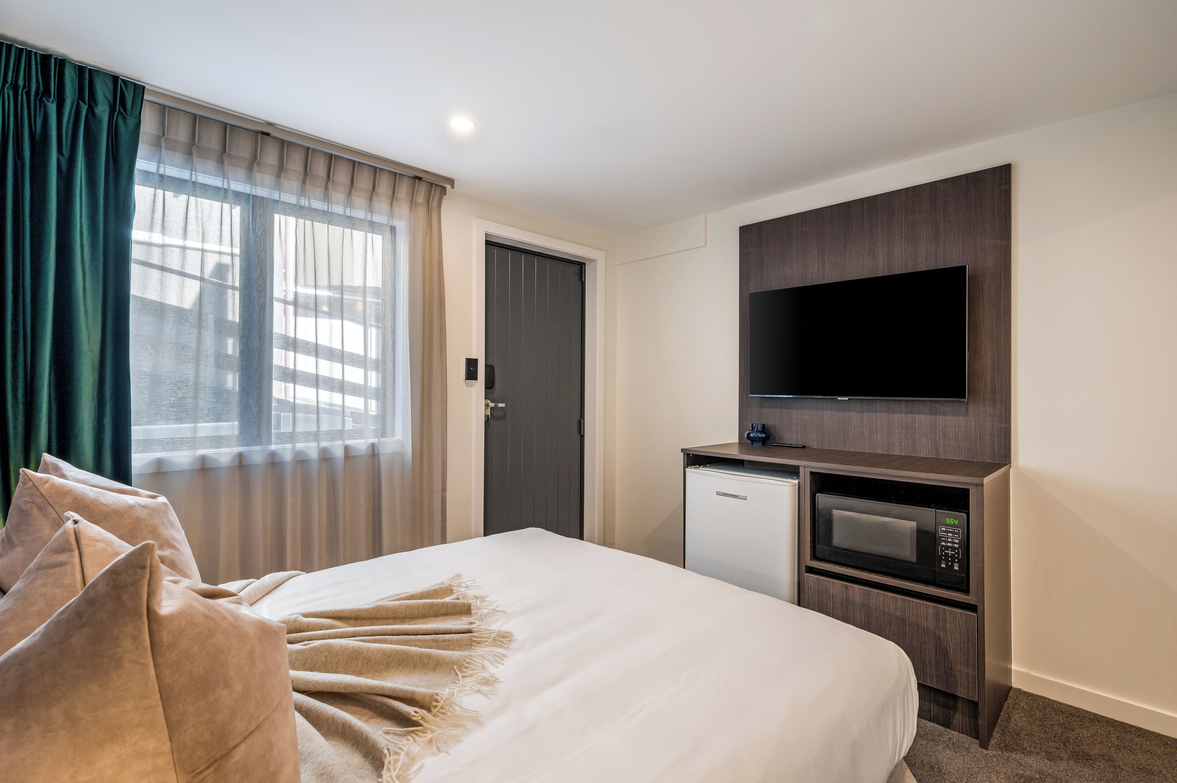 A hotel room with a bed, a flat-screen TV mounted on a wooden panel, a mini-fridge, and a microwave.