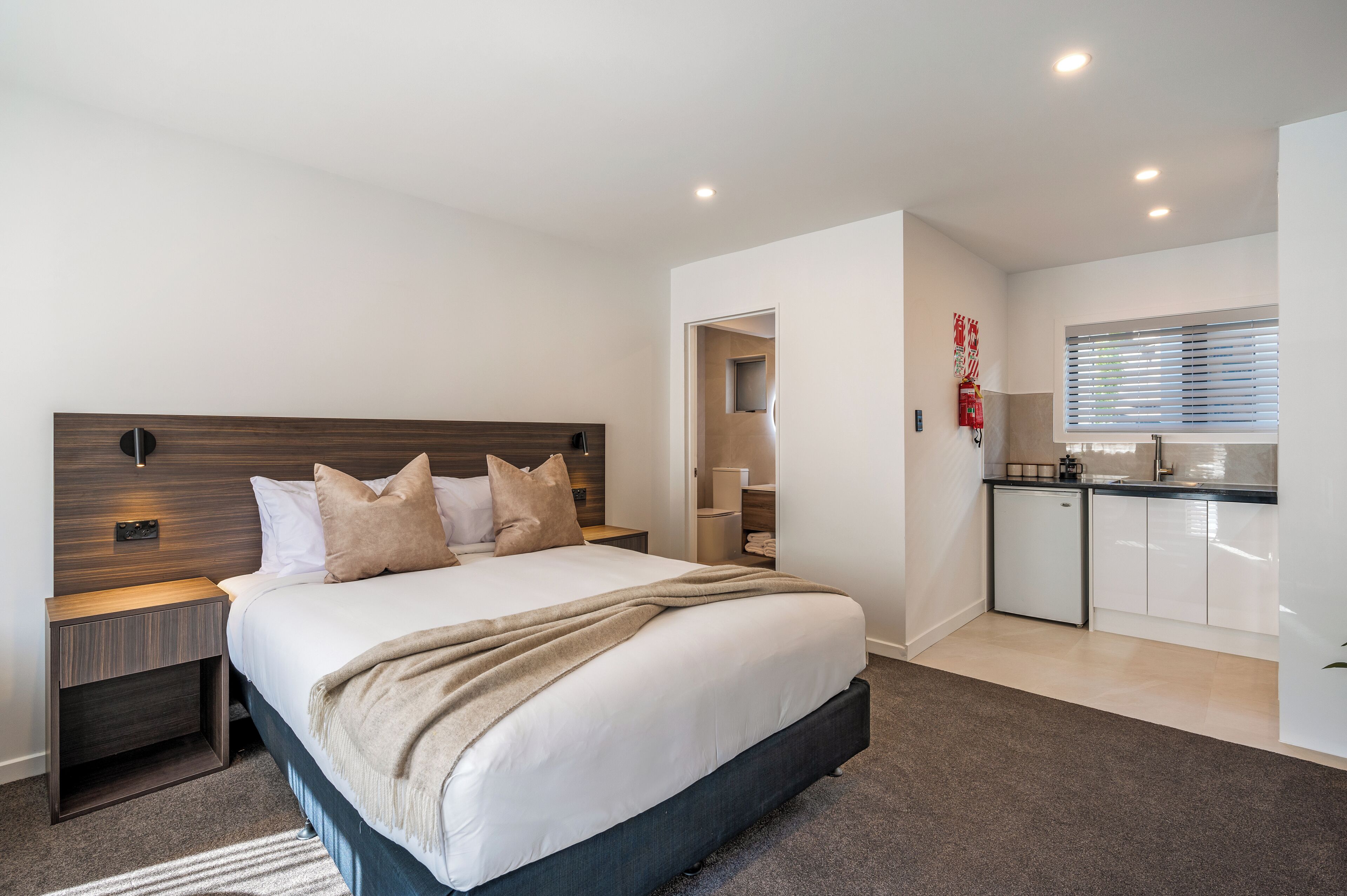 A modern hotel room with a large bed, a small bedside table, a built-in desk, and a kitchenette.