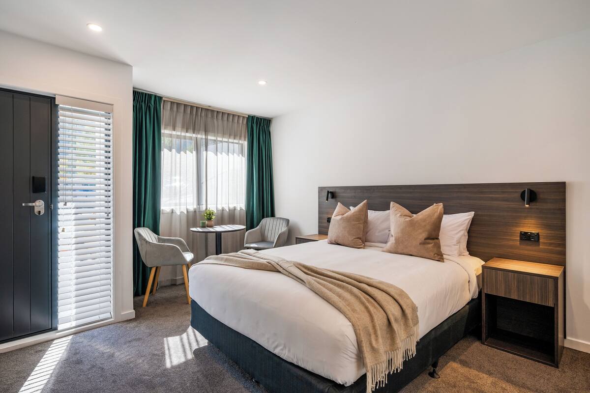 Una habitación de hotel moderna con una cama grande, una mesita de noche, una silla, una pequeña mesa redonda y una puerta con persianas.