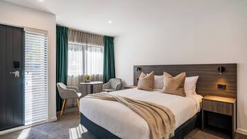 A modern hotel room with a large bed, a bedside table, a chair, a small round table, and a door with blinds.