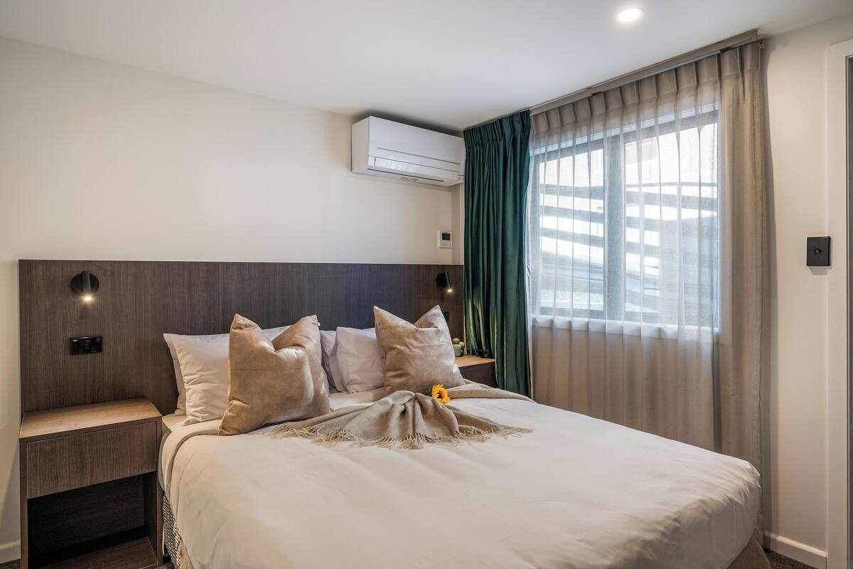 Una cama bien hecha con una manta beige y almohadas, una mesita de noche con una lámpara, un aire acondicionado de pared y una ventana con cortinas traslúcidas.