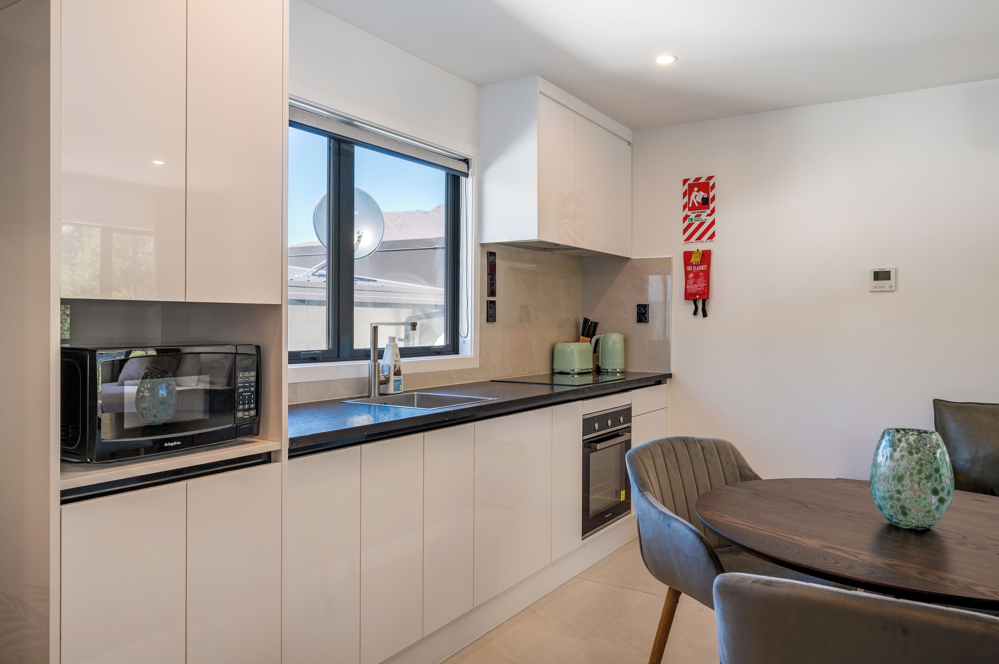 A modern kitchen with white cabinets, a black microwave, a sink, and a window. There is a dining table with chairs and a decorative vase.