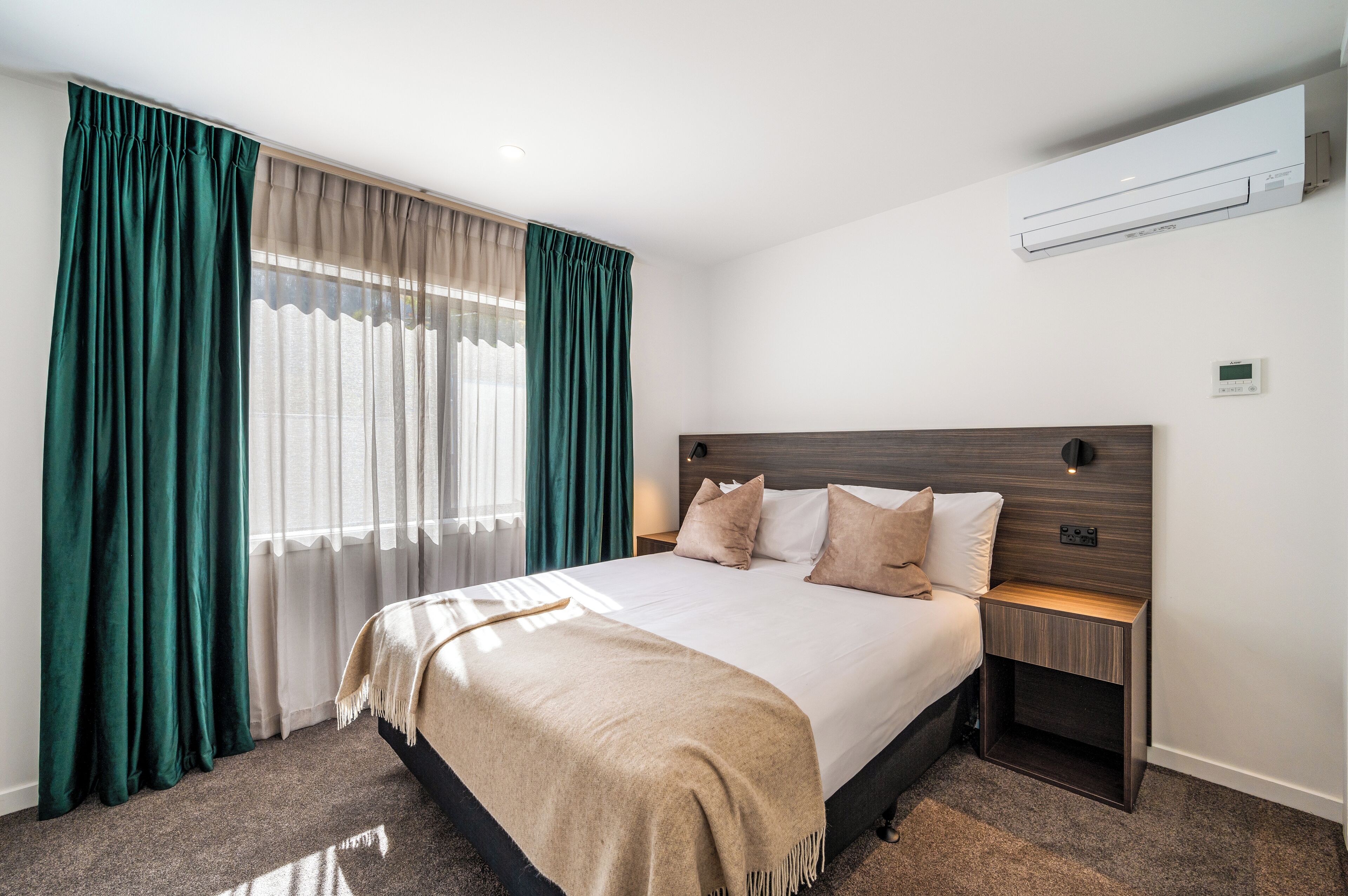 A hotel room with a large bed, a wooden headboard, a nightstand, and a wall-mounted air conditioner.