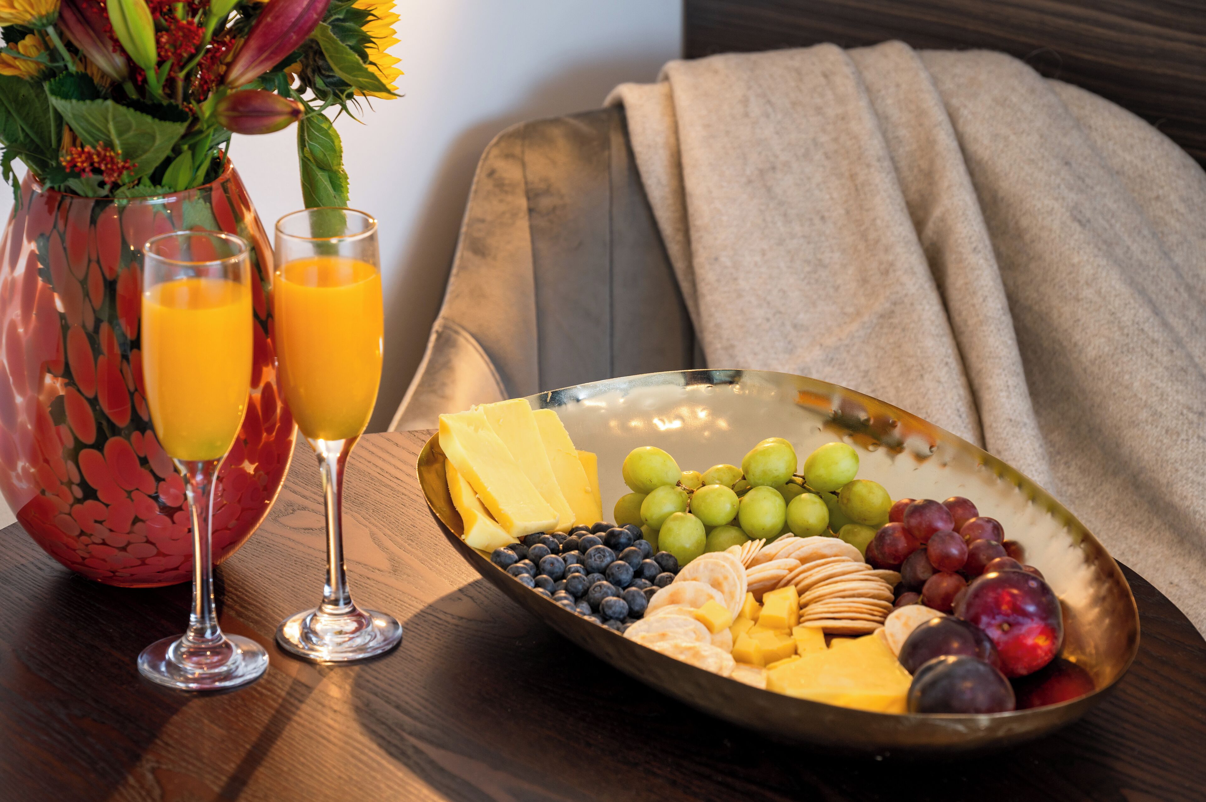 A breakfast setup with a fruit and cheese platter, two glasses of orange juice, and a vase with flowers.