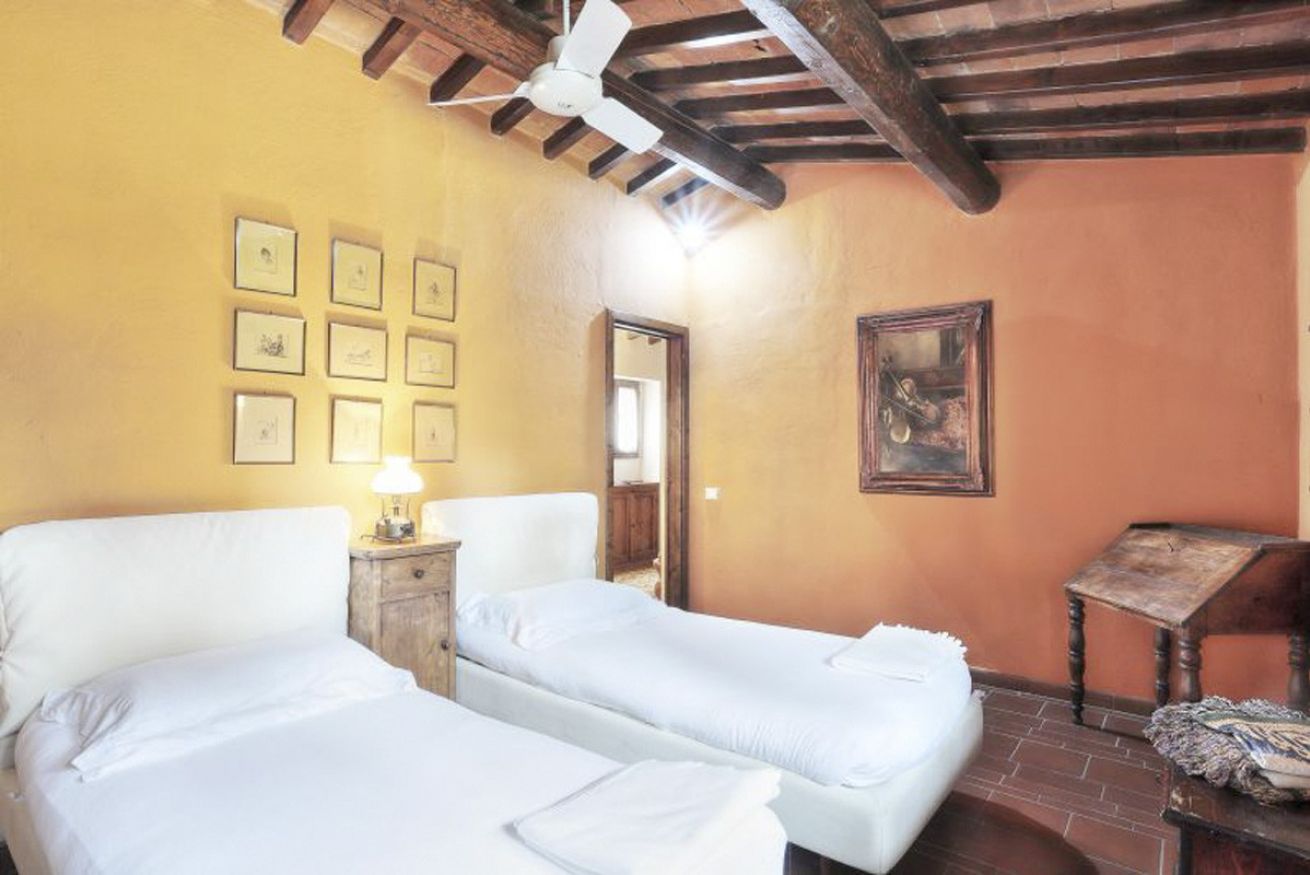 A room with two beds, a ceiling fan, a wooden table, and a wall with framed pictures.