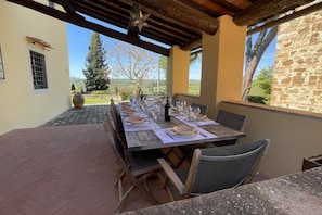 Un área de comedor al aire libre con una larga mesa de madera dispuesta para una comida, rodeada de muros de piedra y vistas al campo.