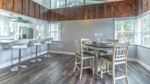 Comedor con techo de madera, cocina al fondo y un ventanal con persianas.