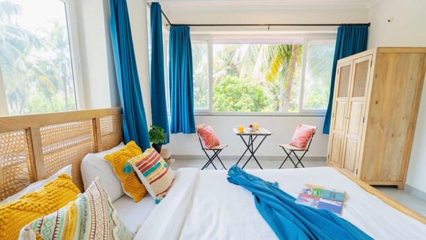 A room with a bed, a wooden wardrobe, a small table with chairs, and a window with curtains. - Mona Collective at Serenova (Calangute)