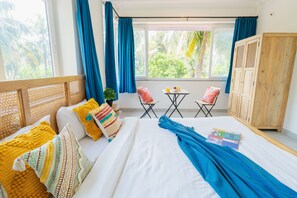 A room with a bed, a wooden wardrobe, a small table with chairs, and a window with curtains. - Mona Collective at Serenova (Calangute)