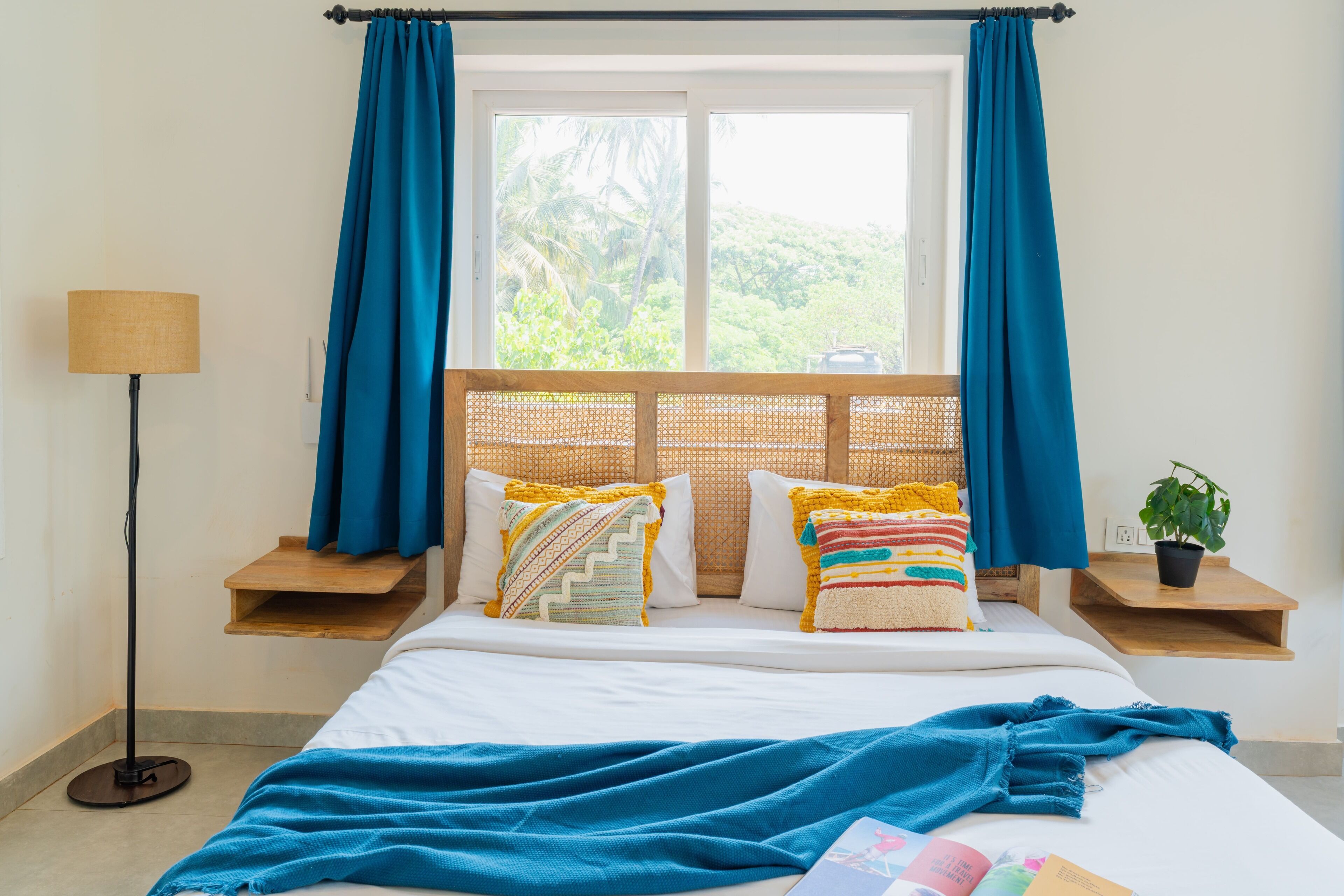 A neatly arranged bedroom with a bed, pillows, a bedside table with a lamp, a window with curtains, and a plant on a small table.