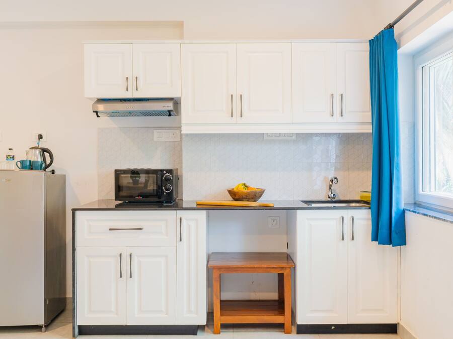 A compact kitchen with white cabinets, a microwave, a kettle, and a small wooden stool.