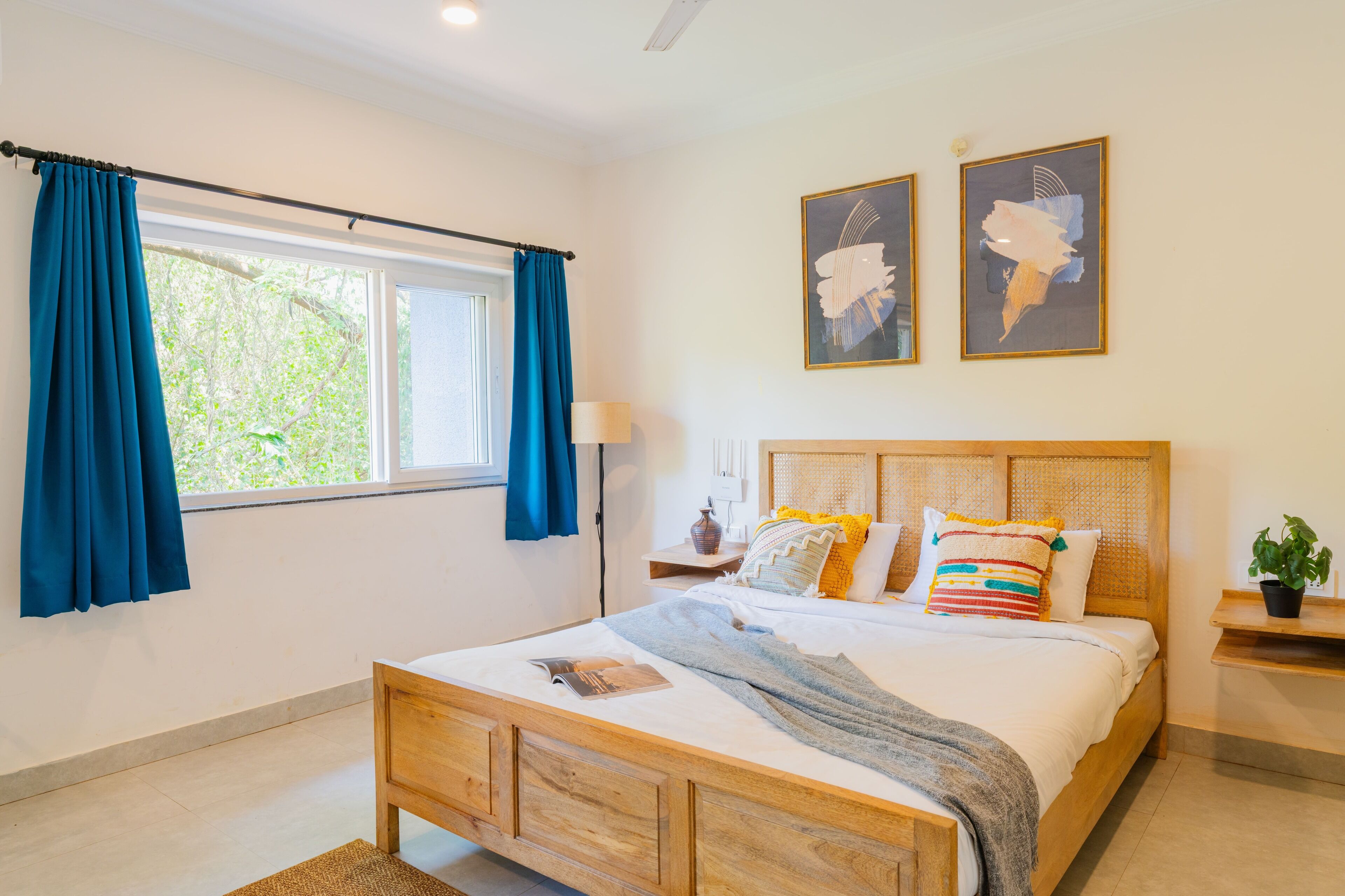 A bedroom with a wooden bed, a window with blue curtains, a bedside table with a lamp, and two framed artworks on the wall.