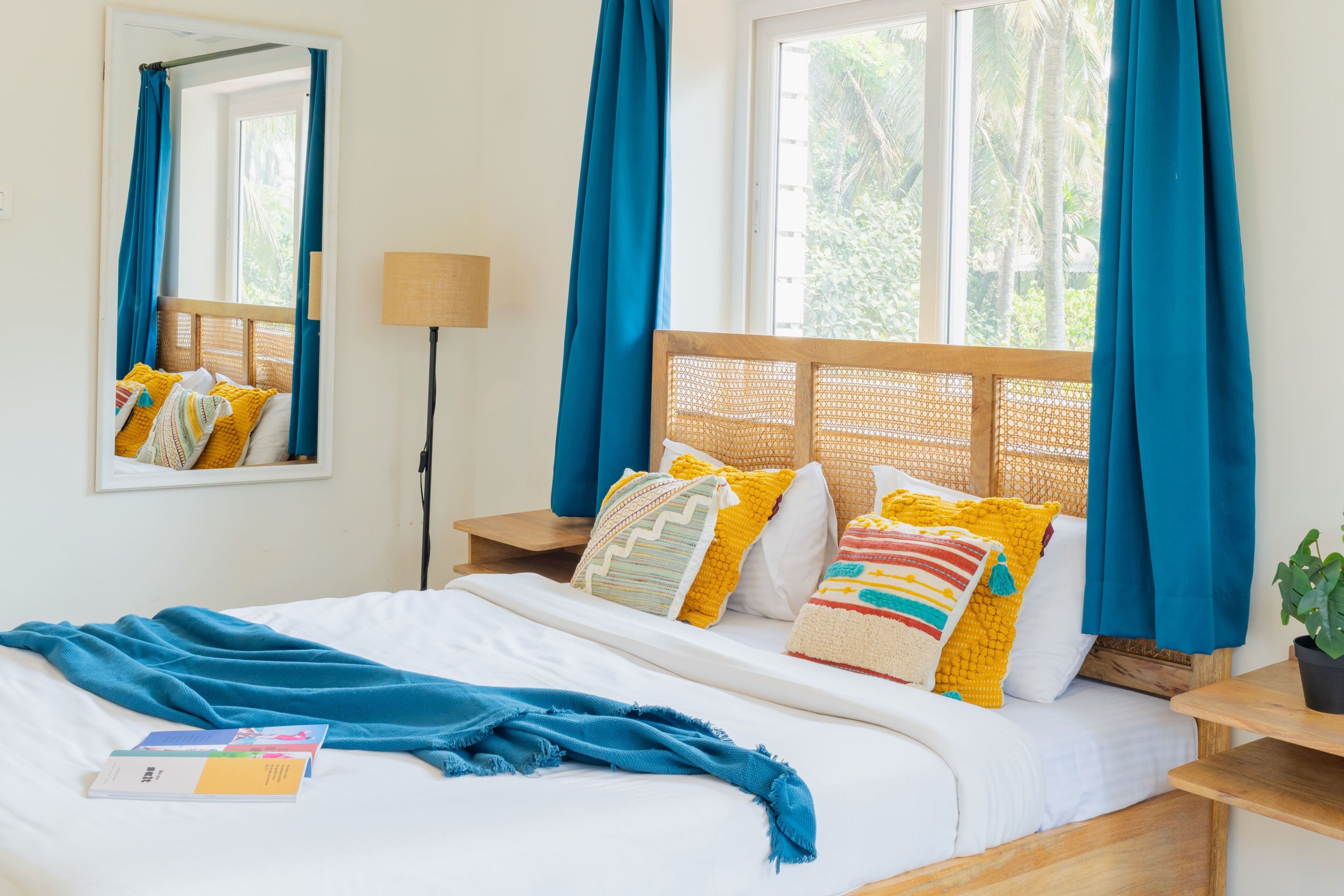 A neatly made bed with a blue blanket, a wooden headboard, and colorful pillows. There is a bedside table with a lamp and a potted plant.