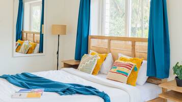 A neatly made bed with a blue blanket, a wooden headboard, and colorful pillows. There is a bedside table with a lamp and a potted plant.
