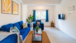 A living room with a blue sofa, a glass coffee table, and a TV mounted on the wall. - Mona Collective at Serenova (Calangute)