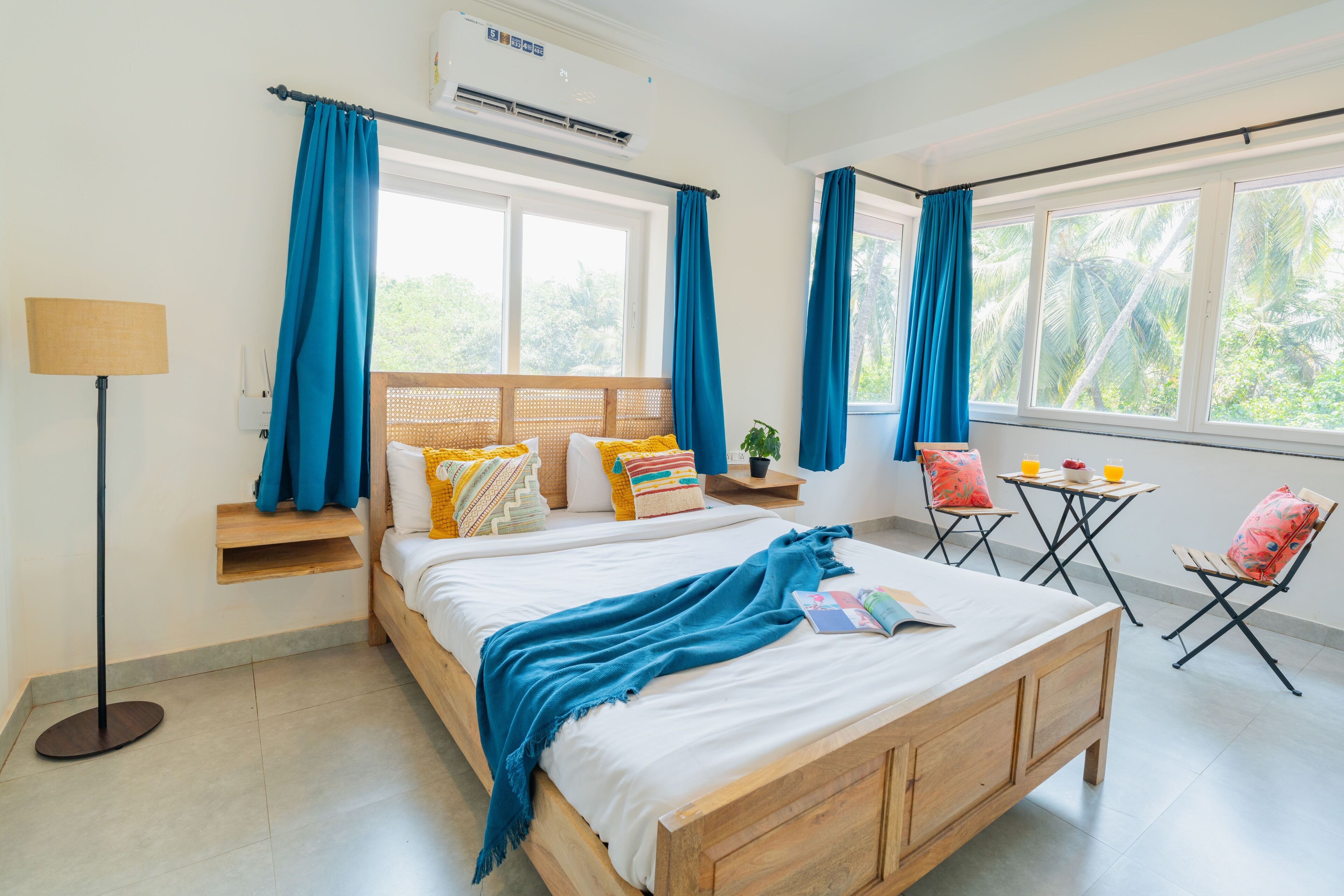 A bedroom with a wooden bed, blue curtains, a bedside table with a lamp, a small table with two chairs, and a view of greenery outside.