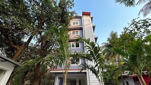 A modern two-story building with a flat roof and a red-tiled roof section. There are trees and a paved area in front of the building. - Mona Collective at Serenova (Calangute)