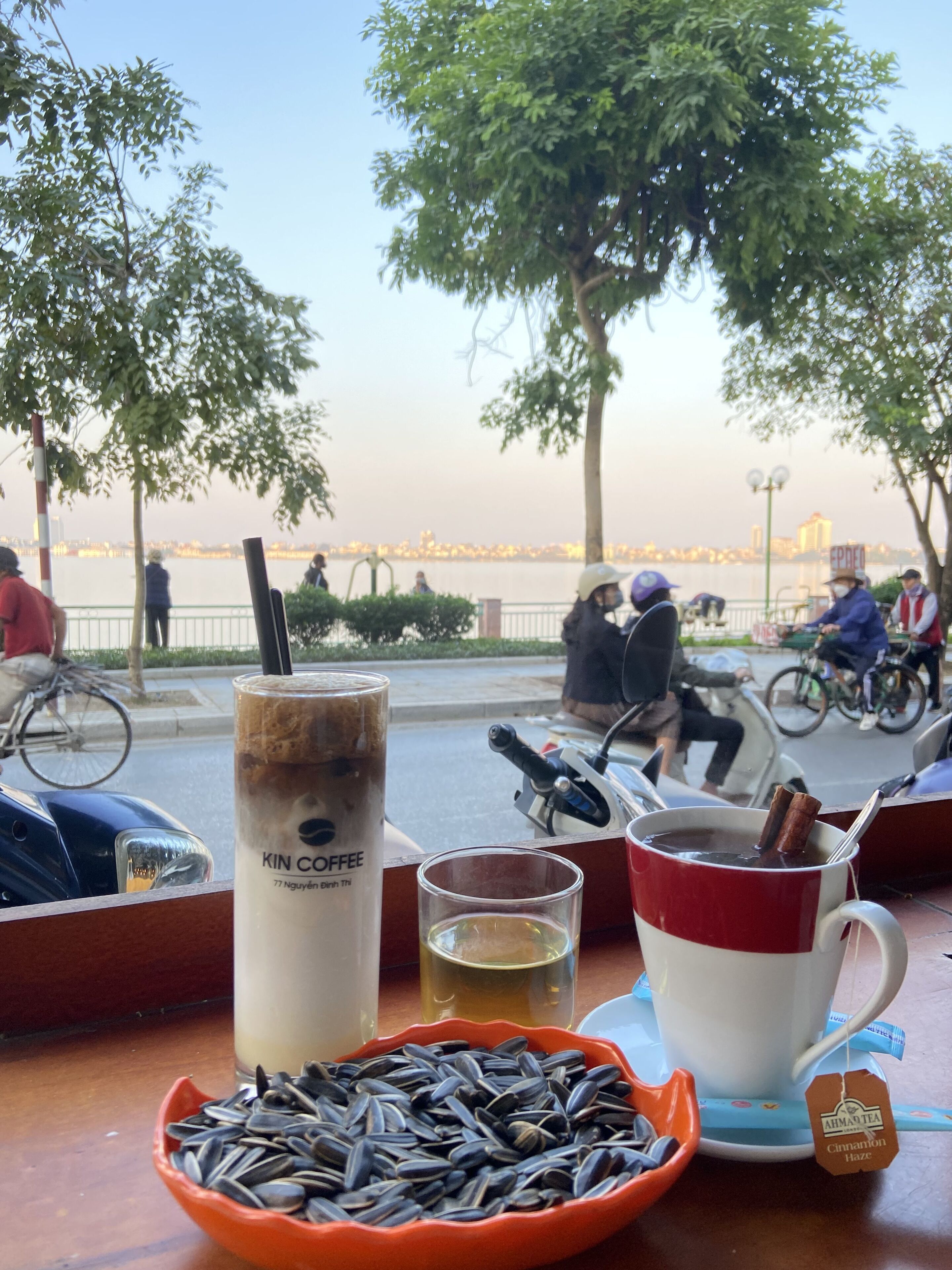 A table with a drink, a bowl of sunflower seeds, and a view of a river with people and bicycles in the background.