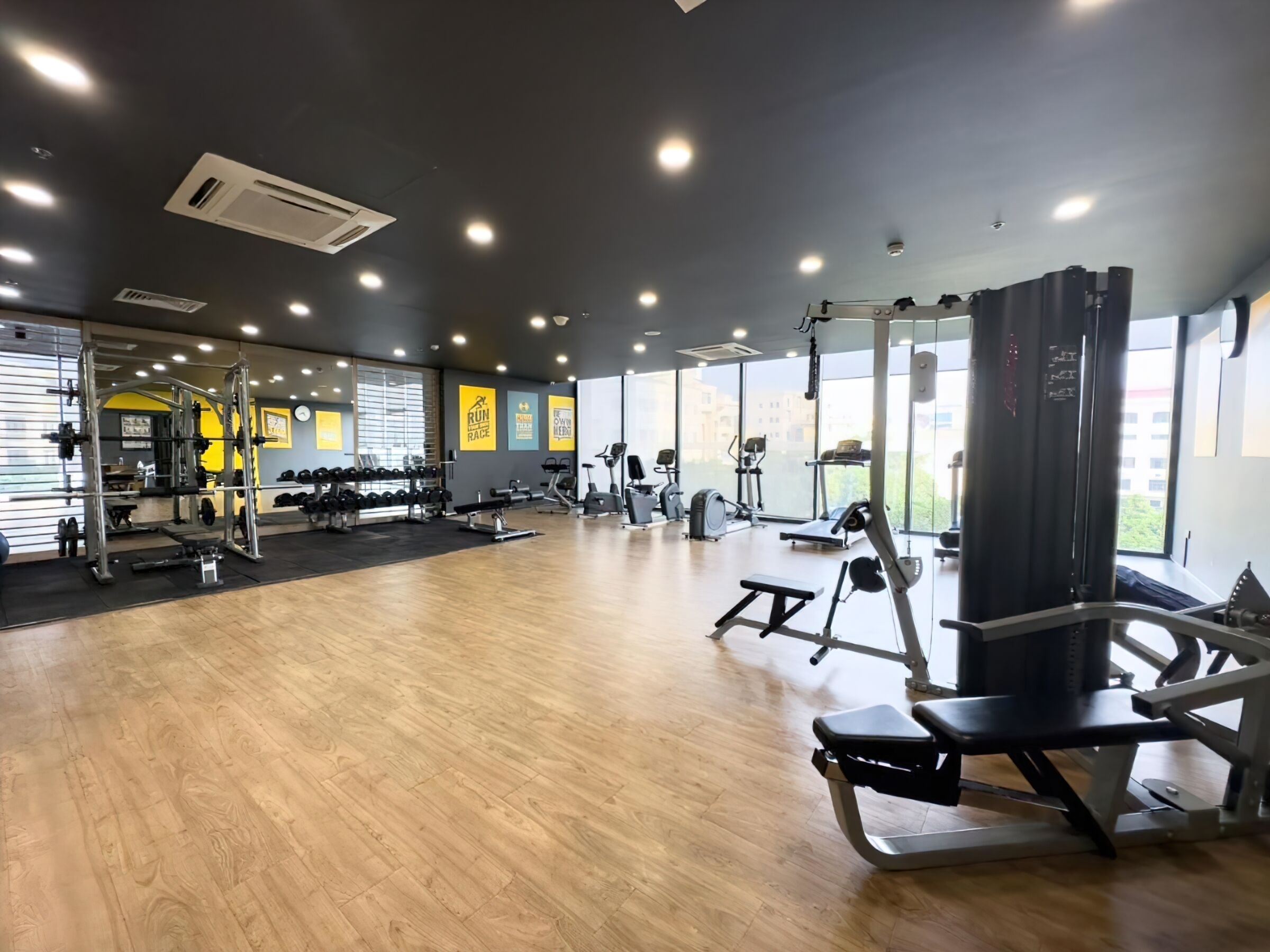 A spacious gym with various exercise equipment, large windows, and a wooden floor.