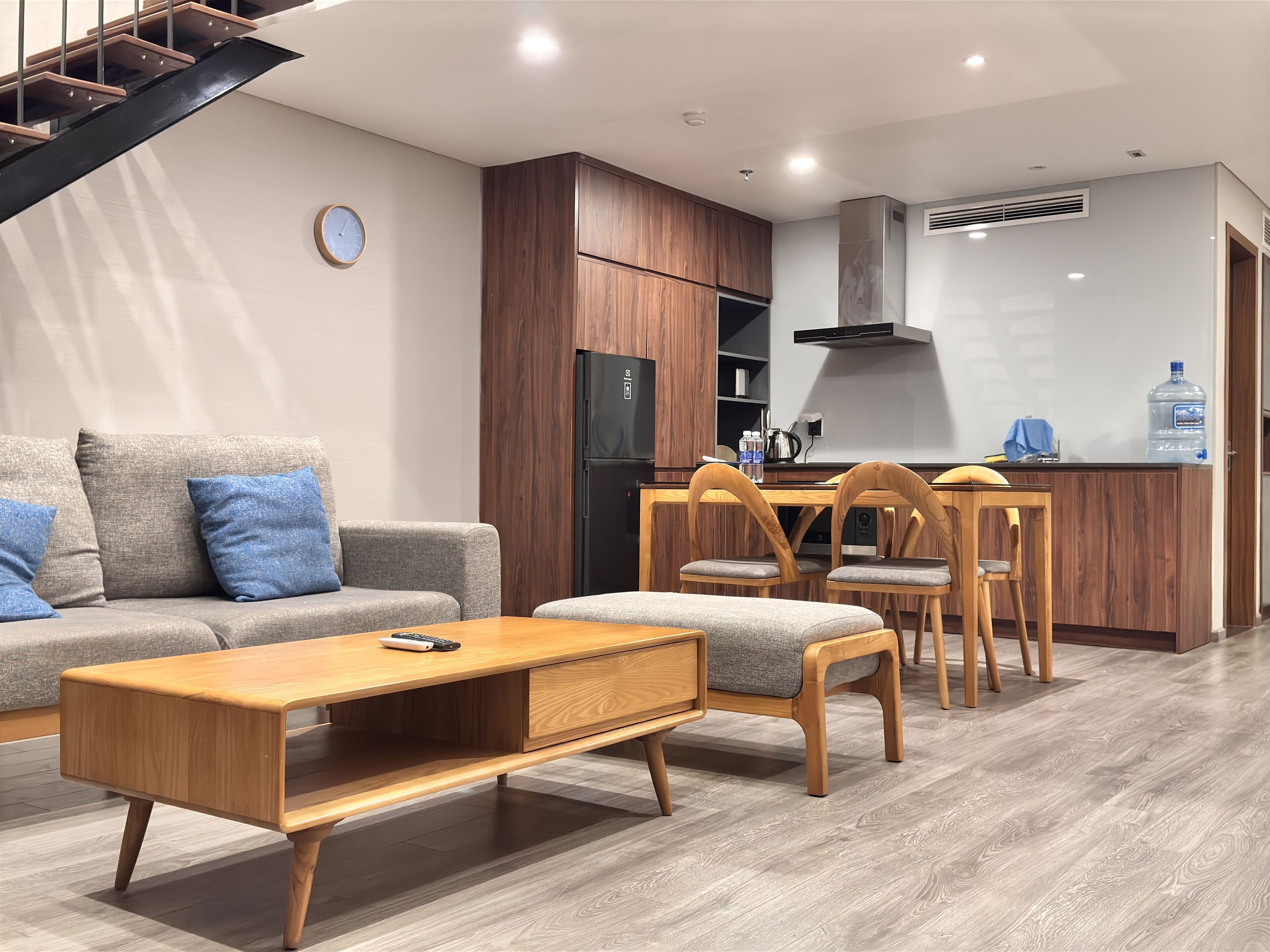 A modern living room with a sofa, coffee table, and wooden chairs. A kitchen area with a refrigerator and dining table is visible in the background.