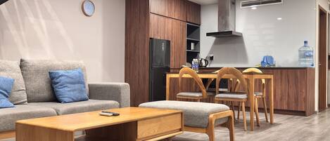 A modern living room with a sofa, coffee table, and wooden chairs. A kitchen area with a refrigerator and dining table is visible in the background.