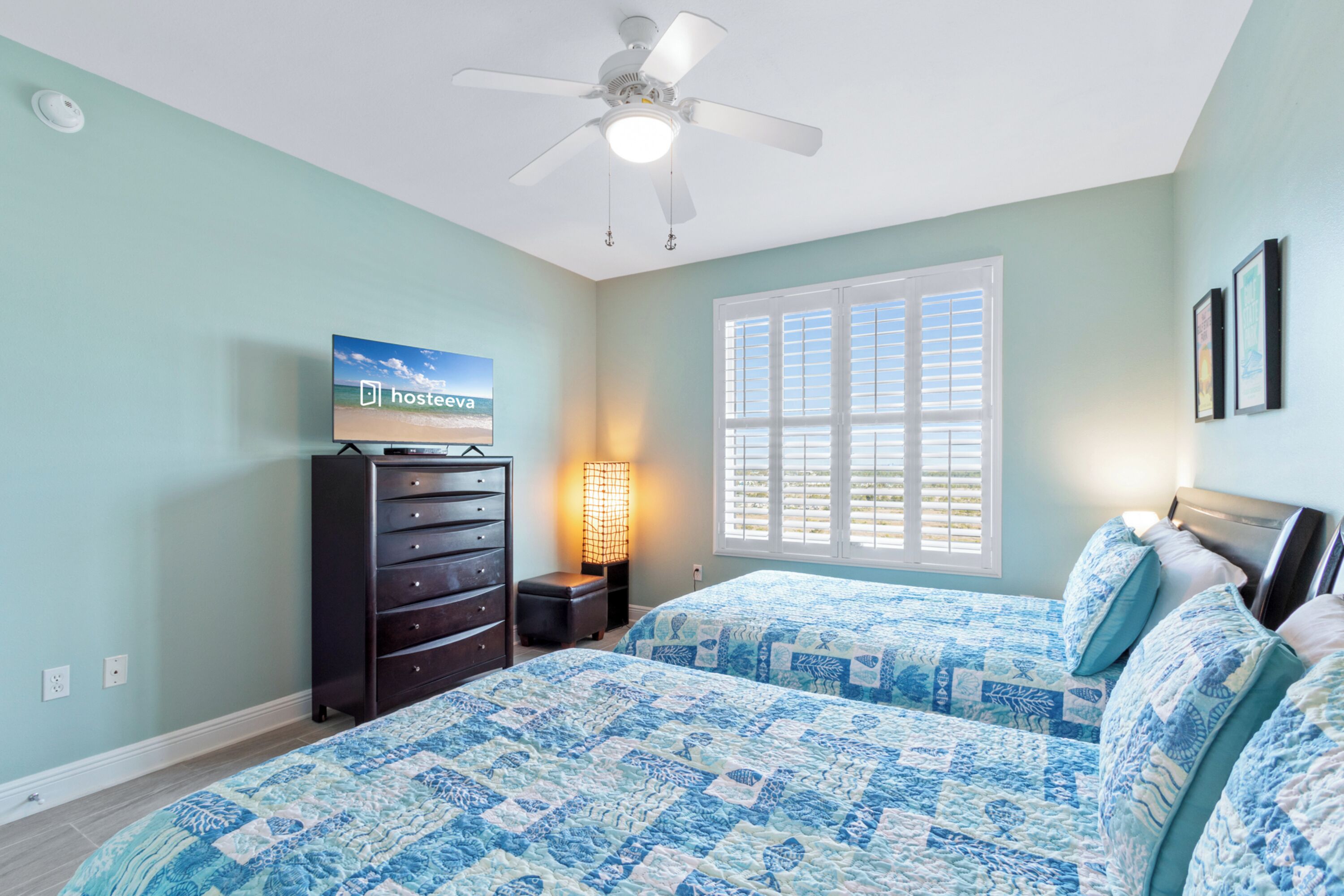A bedroom with two beds, a ceiling fan, a TV on a dresser, and a window with shutters.