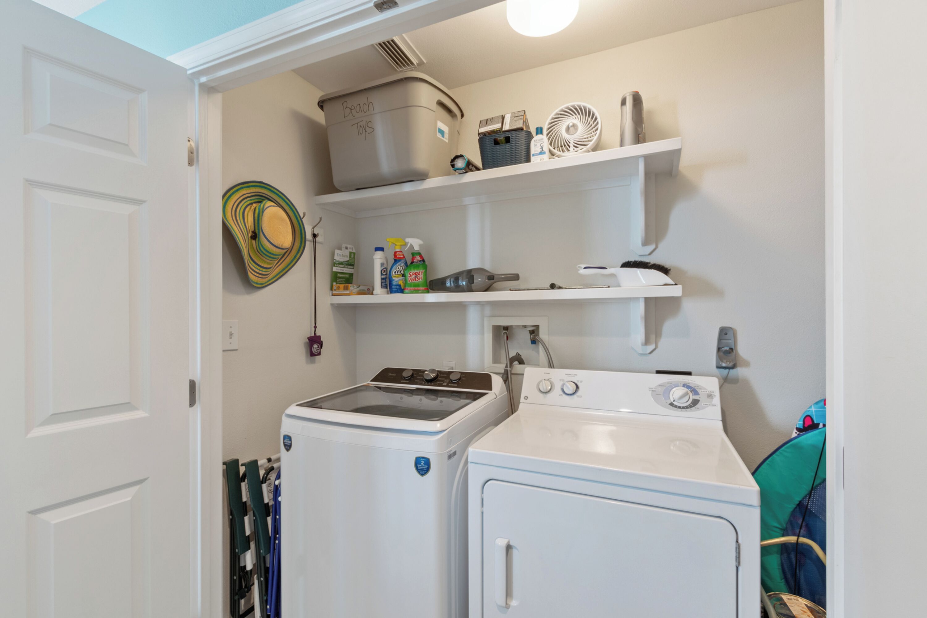 A laundry room with two washers and dryers, shelves with cleaning supplies, and a basket labeled "Beach Toys".
