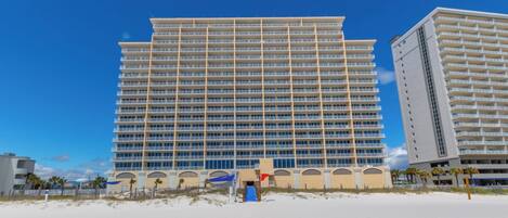 Un hotel de altura con varios balcones, una playa en primer plano y un cielo azul despejado.