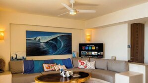 Una sala de estar con un cuadro grande, un televisor de pantalla plana, un ventilador de techo y un sofá con cojines decorativos.