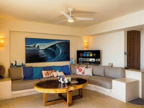 A living room with a large painting, a flat-screen TV, a ceiling fan, and a sofa with decorative pillows. - Luxury Penthouse at Aura del Mar (Zihuatanejo)