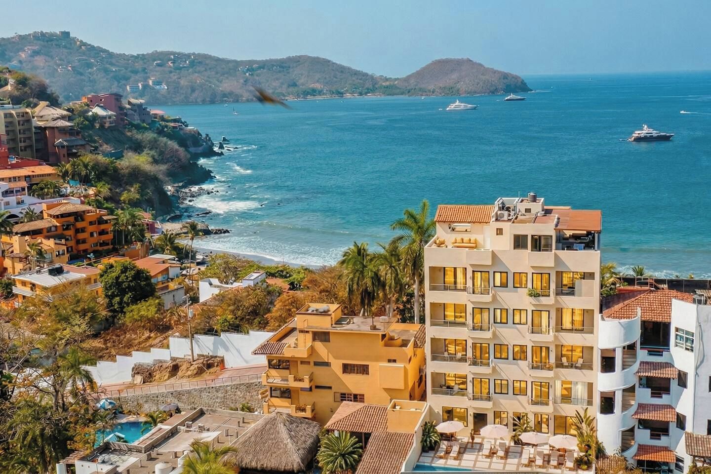 A coastal hotel with multiple balconies, a swimming pool, and a view of the sea and hills.