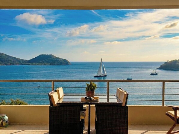 A balcony with wicker furniture, a potted plant, and a view of the sea with sailboats.