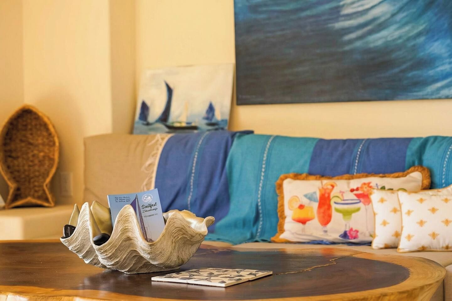 A room with a wooden coffee table, a card game, a decorative shell, a sailboat painting, and a sofa with patterned cushions.