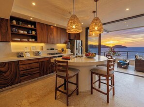 A modern kitchen with wooden cabinets, a central island with bar stools, and a view of the ocean at sunset.