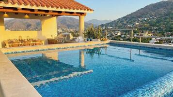 A swimming pool with a tiled edge, a pergola with a tiled roof, and a view of a hilly landscape in the background.