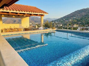 A swimming pool with a tiled edge, a pergola with a tiled roof, and a view of a hilly landscape in the background.