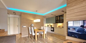 Un salon moderne comprenant un coin repas, un plancher en bois, un téléviseur à écran plat et une cuisine équipée d’appareils électroménagers intégrés.