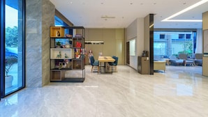 Eine moderne Hotellobby mit Marmorboden, einer zentralen Sitzecke mit Tisch und Stühlen sowie einem großen Bücherregal voller verschiedener Gegenstände.