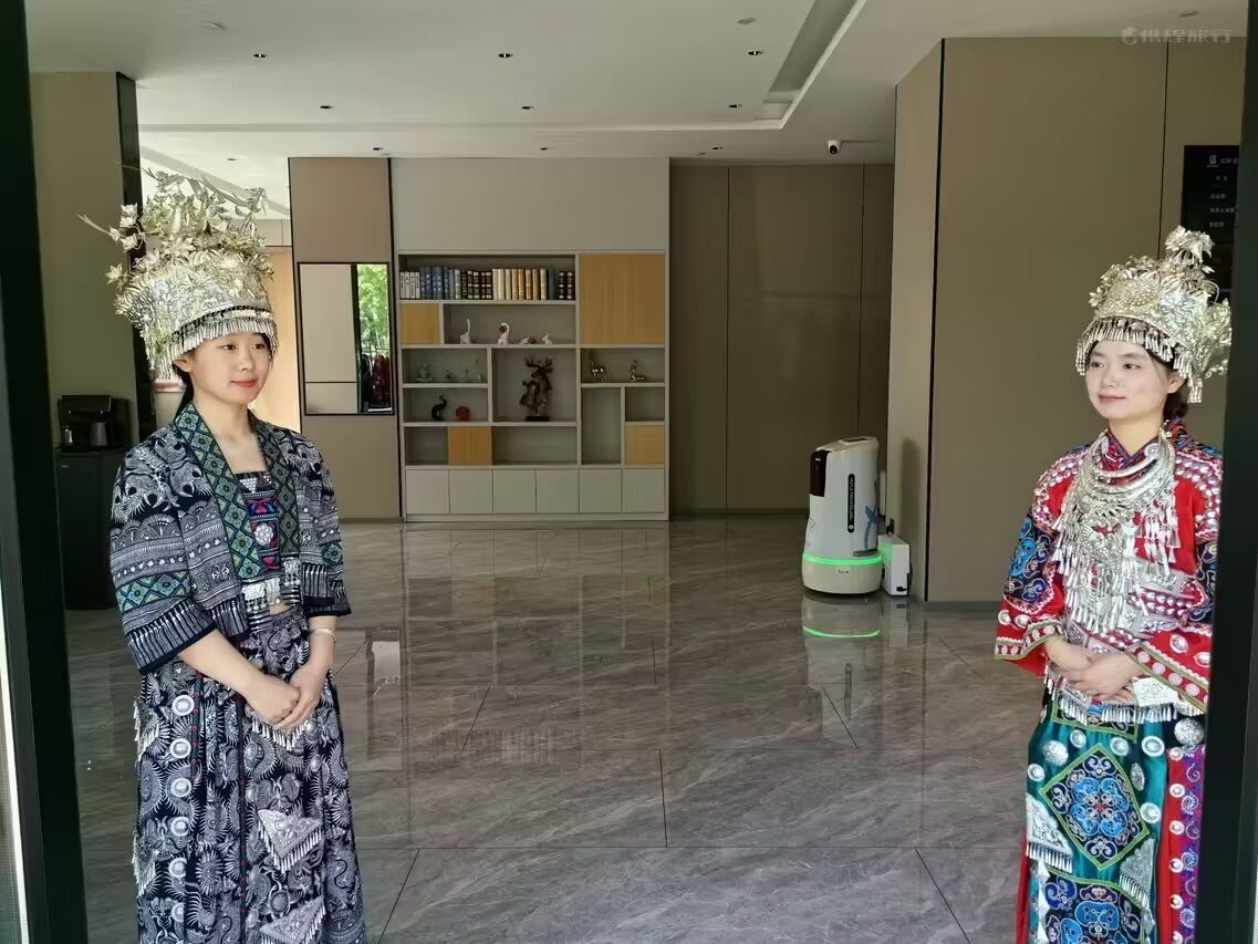 Two individuals in traditional attire standing in a modern interior space with a polished floor, a bookshelf, and a coffee machine.