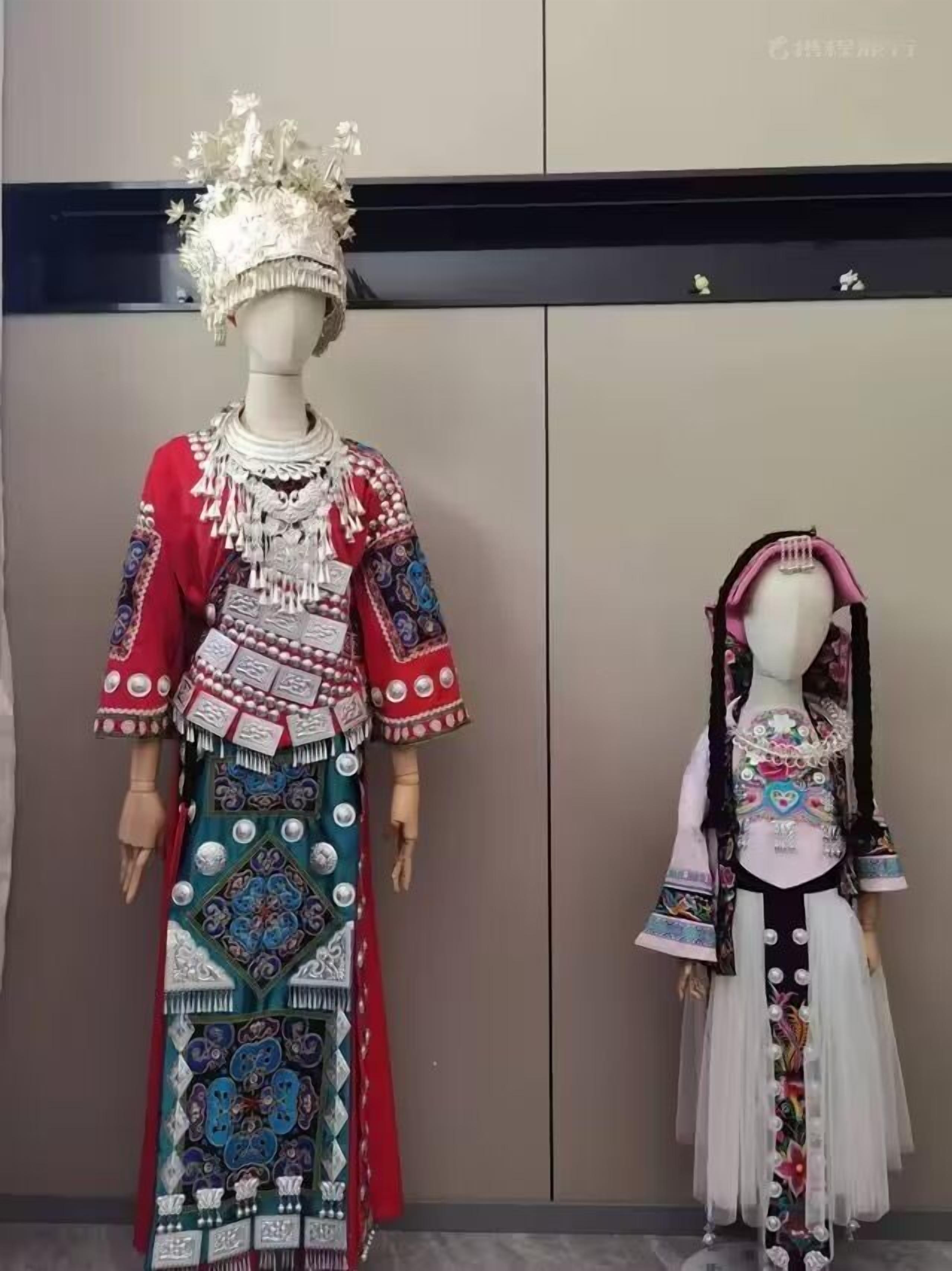 Zwei Schaufensterpuppen, bekleidet mit traditioneller Kleidung und aufwendigen Kopfschmuck sowie Schmuck, vor einer schlichten Wand.