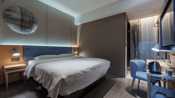 Ein modernes Hotelzimmer mit einem großen Bett, einem Schreibtisch mit Stuhl, einem Fernseher und einem einzigartigen Kunstwerk an der Wand.