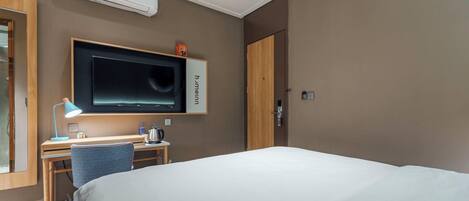 A hotel room with a bed, a desk with a chair, a television, and an air conditioning unit.