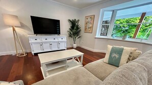 Uma sala de estar com televisão de tela plana, um móvel de armazenamento branco, uma mesa de centro, um sofá bege com almofada de folha de palmeira e uma janela grande com vista para áreas verdes.