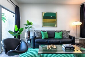 Un soggiorno moderno con un divano in pelle nera, cuscini verdi, un tavolino da caffè in vetro con libri, una poltrona nera e un grande quadro astratto appeso alla parete.