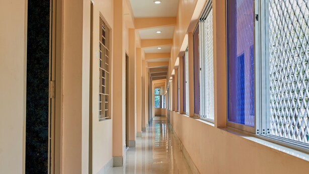 A long, well-lit corridor with a tiled floor and beige walls, leading to a room with a large window and a metal grille. - Deb Guest House (Barakpur)
