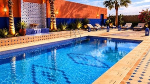 Una piscina con bordes de azulejos, sillas de descanso, palmeras y un edificio con paredes ornamentadas.