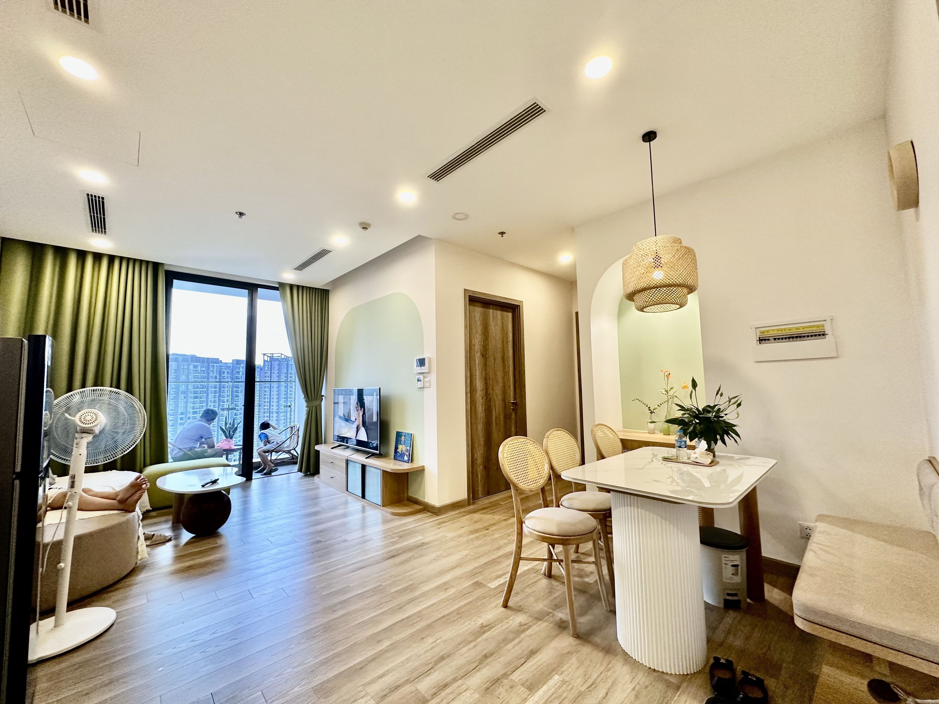 A modern living room with a dining area, wooden flooring, a large window with curtains, a ceiling fan, and a TV.