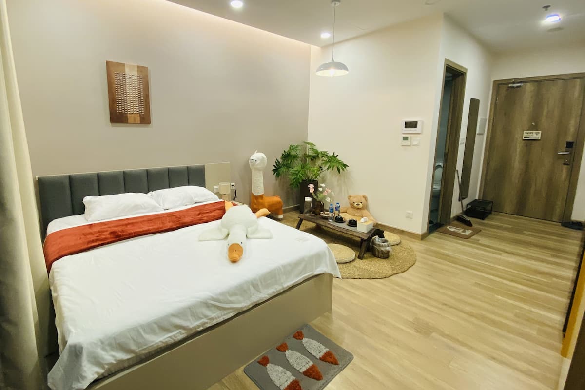Quarto de hotel moderno com uma cama grande, uma mesa de cabeceira com luminária, um vaso de planta e um ursinho de pelúcia.