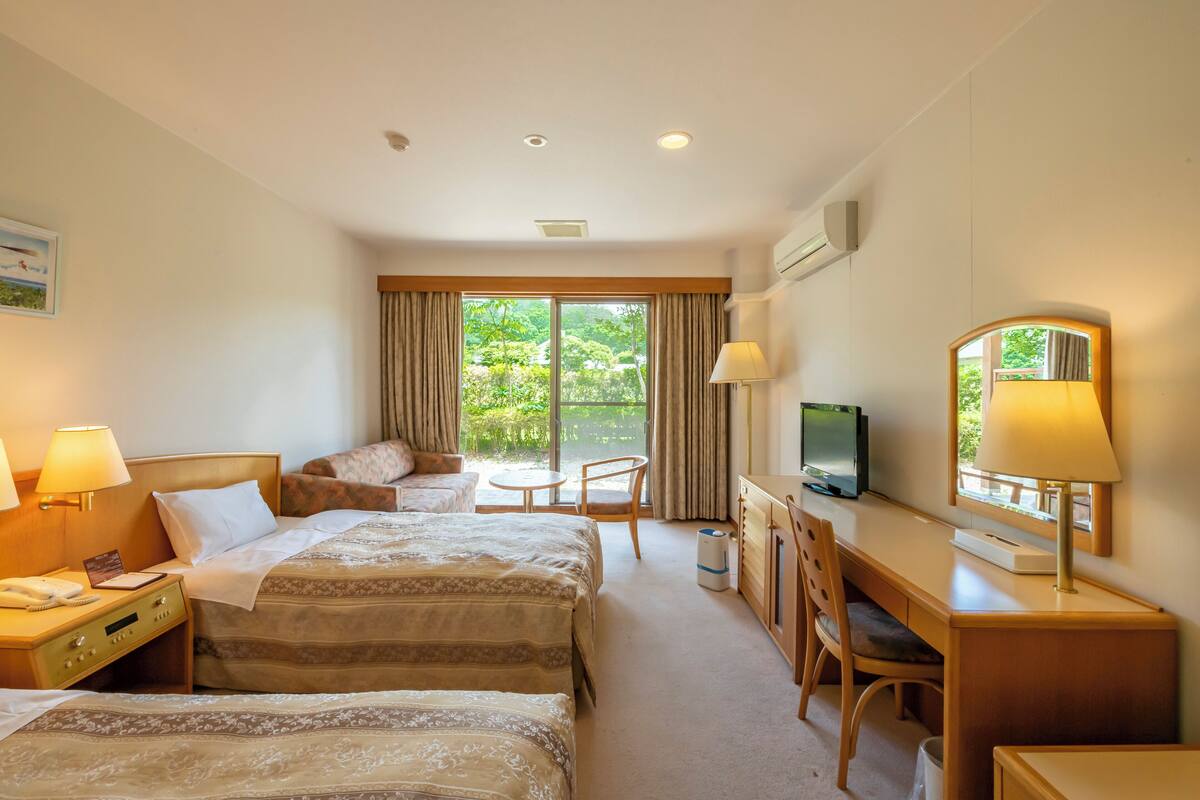 Quarto de hotel com duas camas, uma escrivaninha, uma cadeira, uma televisão e uma janela com cortinas.
